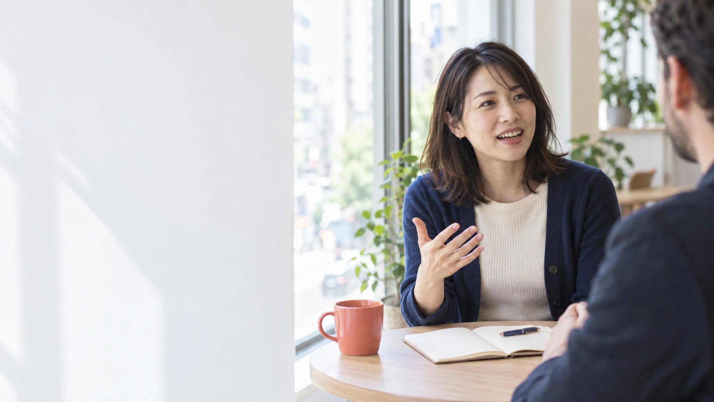 窓辺の木のテーブルで、先生に英語で話す女性。手ぶりをまじえ、言葉を探しながら笑っている（写真は見本）