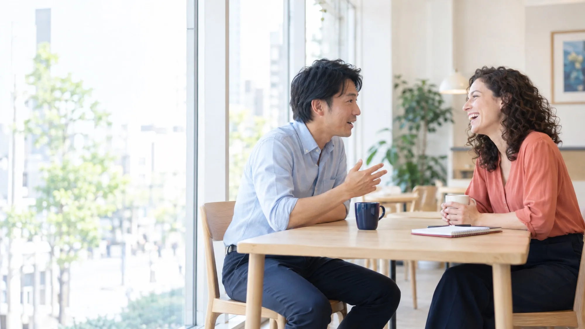 窓辺のテーブルで、男性が英語で話し、向かいの先生が声を出して笑っている（写真は見本）